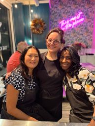 Three smiling women in black aprons pose together inside a bright craft studio with a pink neon sign, patterned wallpaper, a decorative wreath and string lights.