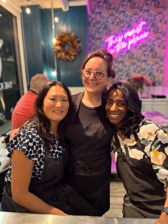 Three smiling women in black aprons pose together inside a bright craft studio with a pink neon sign, patterned wallpaper, a decorative wreath and string lights.