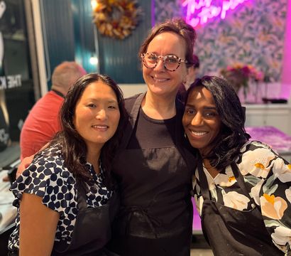 Three smiling women in black aprons pose together inside a bright craft studio with a pink neon sign, patterned wallpaper, a decorative wreath and string lights.