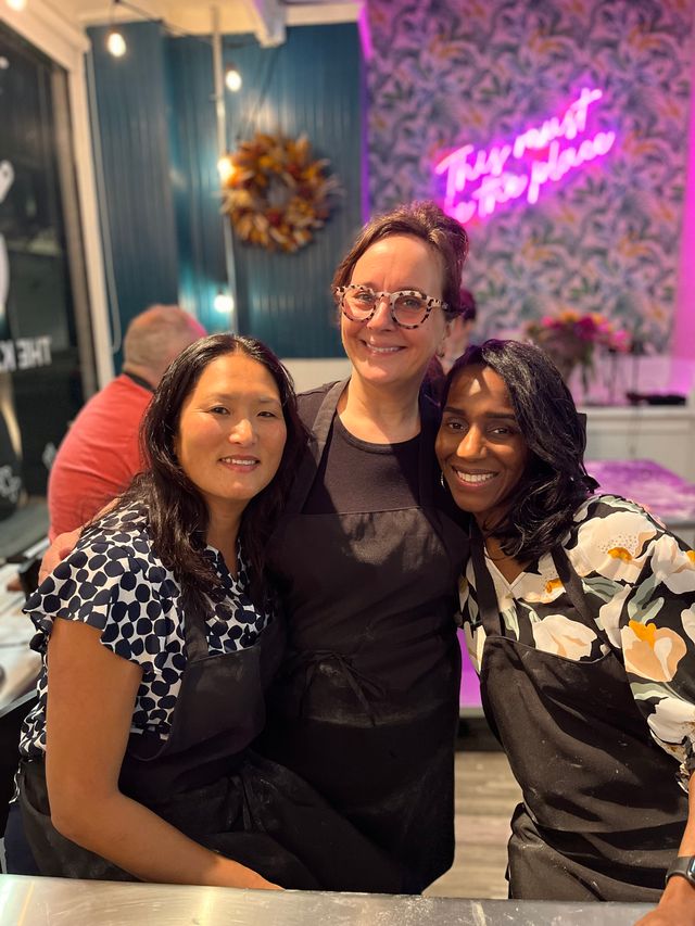 Three smiling women in black aprons pose together inside a bright craft studio with a pink neon sign, patterned wallpaper, a decorative wreath and string lights.