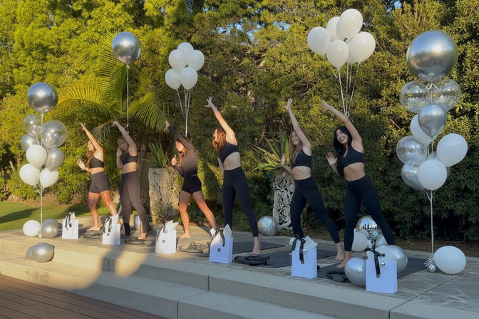 Six participants in black activewear performing synchronized side stretches on yoga mats on a sunlit outdoor patio, surrounded by silver and white balloons, gift bags, and a lush green garden backdrop — a celebratory outdoor fitness scene at golden hour.