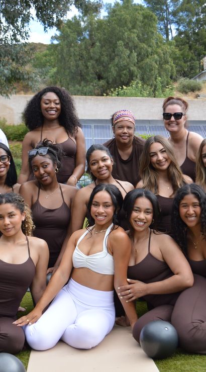 Smiling group of women in matching brown workout sets (one in white) posing on yoga mats with medicine balls at an outdoor wellness/fitness retreat on a grassy hillside with trees and solar panels in the background.