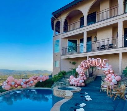 Hilltop luxury villa outdoor pool with stone spa and sweeping mountain views, decorated for a bridal celebration with rose-gold 'BRIDE' balloon arch and pink balloon garlands along the railing under a clear blue sky