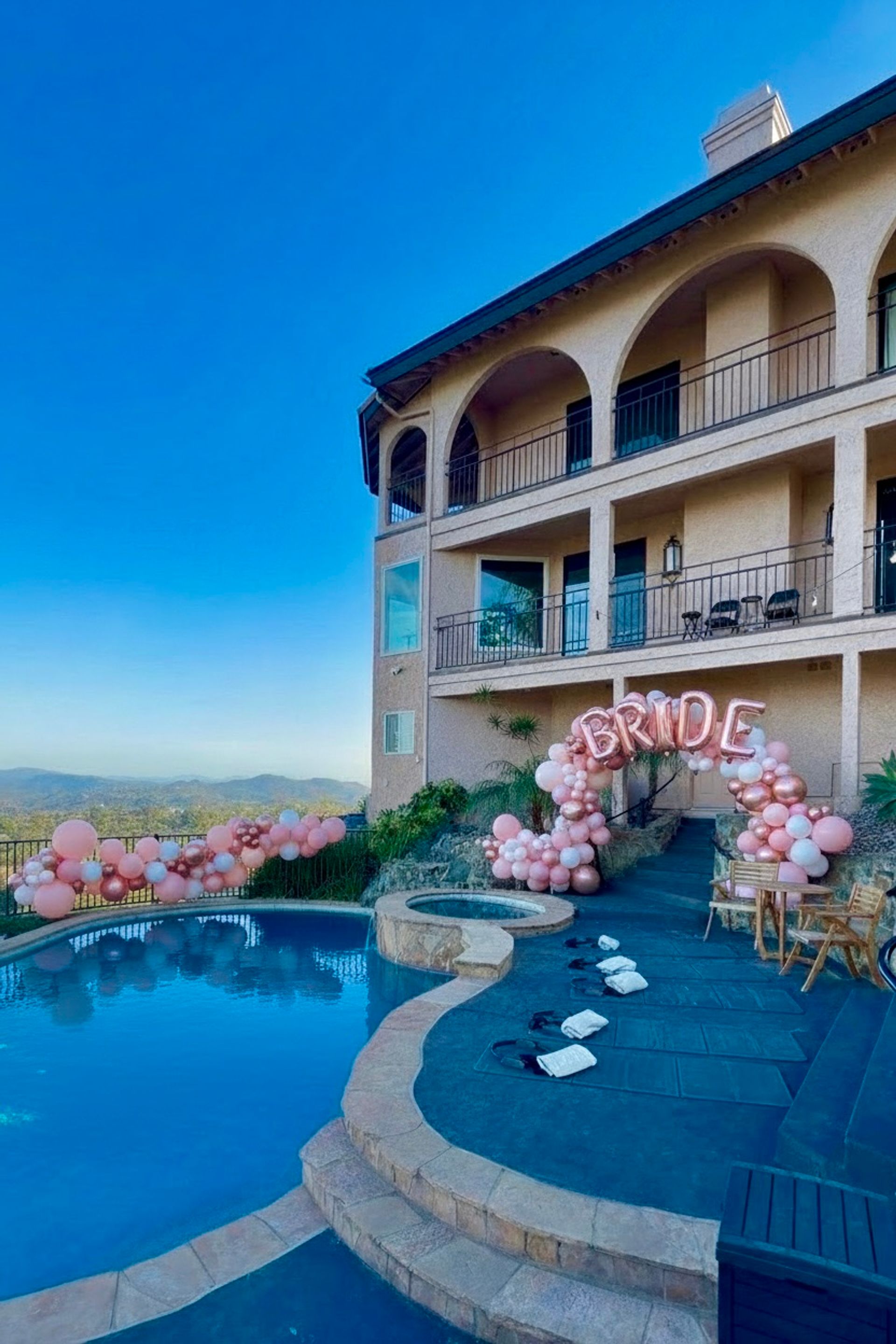 Hilltop luxury villa outdoor pool with stone spa and sweeping mountain views, decorated for a bridal celebration with rose-gold 'BRIDE' balloon arch and pink balloon garlands along the railing under a clear blue sky