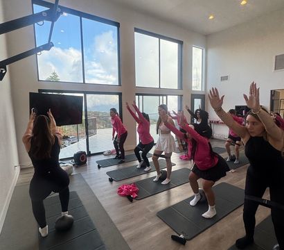 High-energy group squat class in a bright studio with floor-to-ceiling windows overlooking rolling hills; participants on mats, instructor up front, pink workout gear and a disco ball on the floor.