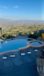 Hillside backyard with a blue infinity-style pool and circular spa on a stone patio, lined with exercise mats and weighted bags, overlooking a sunlit valley and distant mountain range under a clear blue sky.