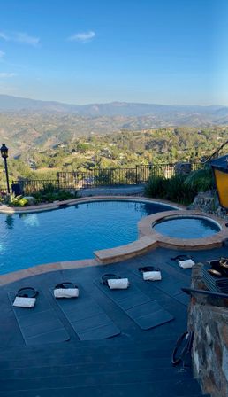 Hillside backyard with a blue infinity-style pool and circular spa on a stone patio, lined with exercise mats and weighted bags, overlooking a sunlit valley and distant mountain range under a clear blue sky.