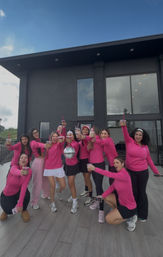 Group of women in matching pink tops toasting iced drinks on a modern rooftop terrace in front of a dark contemporary house, smiling and holding a small disco ball.