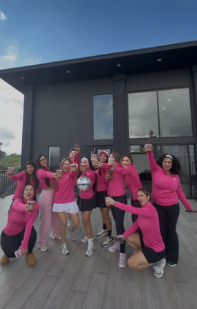 Group of women in matching pink tops toasting iced drinks on a modern rooftop terrace in front of a dark contemporary house, smiling and holding a small disco ball.