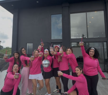 Group of women in matching pink tops toasting iced drinks on a modern rooftop terrace in front of a dark contemporary house, smiling and holding a small disco ball.