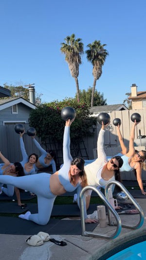 Women in matching light-blue activewear doing side-plank medicine-ball workout next to a backyard pool with palm trees and clear blue sky