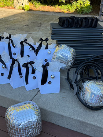 Outdoor fitness setup on a stone patio: white gift bags with black bows and mini disco-ball ornaments, stacked exercise mats, Pilates rings and mirrored disco-cube props ready for a stylish workout event