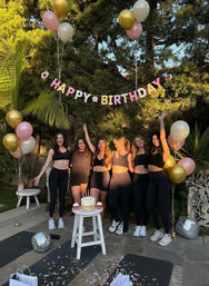 Six women in black workout outfits posing and smiling under a "Happy Birthday" banner in a sunny backyard garden patio with pink and gold balloons, a cake on a stool and disco balls.