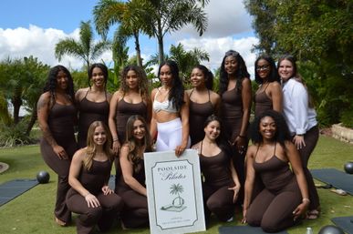 Smiling group of women in brown workout outfits posing on a grassy lawn with palm trees, holding a pilates sign with exercise mats nearby.
