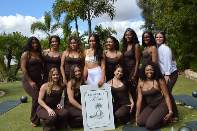 Smiling group of women in brown workout outfits posing on a grassy lawn with palm trees, holding a pilates sign with exercise mats nearby.