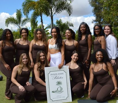 Smiling group of women in brown workout outfits posing on a grassy lawn with palm trees, holding a pilates sign with exercise mats nearby.