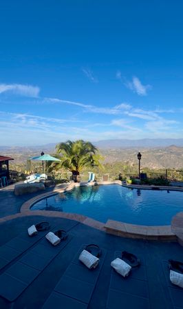 Sunny hilltop pool with a palm tree, lounge mats and towels, turquoise umbrella, and sweeping mountain-and-valley views under a bright blue sky.