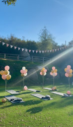 Outdoor yoga setup on a green lawn with black mats, pastel pink and gold balloon bouquets, bunting and a sunbeam under a clear blue sky