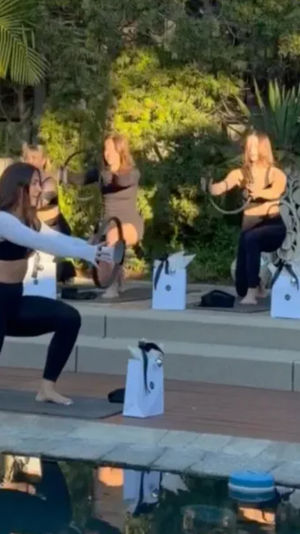 Sunlit outdoor poolside Pilates group: women on mats doing lunges and using Pilates rings beside a backyard pool
