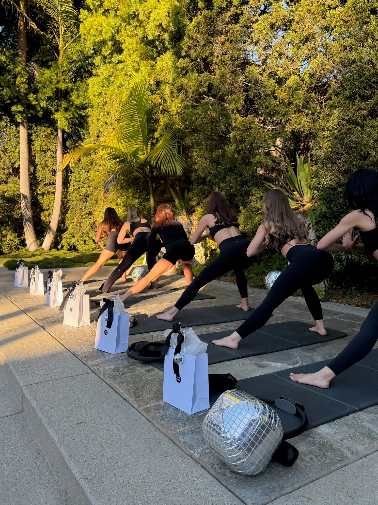 Sunlit backyard fitness class at golden hour — women in black activewear doing lunges on yoga mats beside palm trees, gift bags and a small mirror ball on a stone patio.