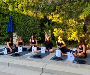 Six women relaxing on yoga mats in a sunlit garden patio at golden hour, holding water bottles with white gift bags nearby.