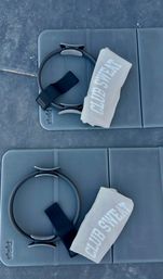 Top-down view of two blue-gray exercise mats, each staged with a Pilates ring, black resistance strap, and a rolled pale towel with bold white lettering, ready for a workout.