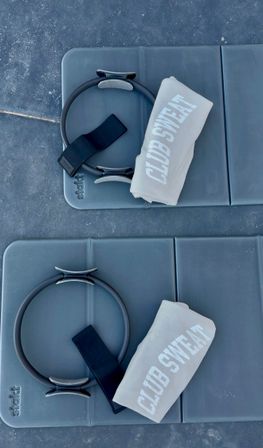 Top-down view of two blue-gray exercise mats, each staged with a Pilates ring, black resistance strap, and a rolled pale towel with bold white lettering, ready for a workout.