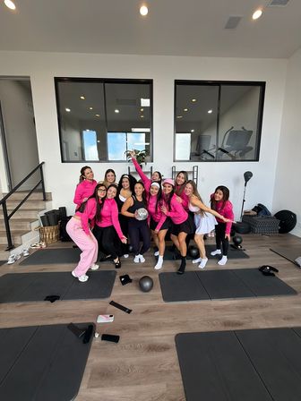 Group of women in pink workout tops posing and laughing in a bright modern fitness studio with exercise mats, small weights, hardwood floor and a silver disco ball.