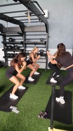 Group fitness class of four women doing squats on black mats in an indoor gym with green turf, kettlebells and medicine balls on wall racks