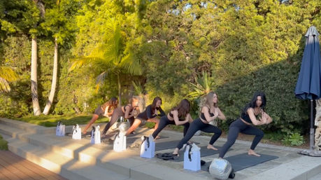 Sunlit outdoor yoga class on a garden patio — a group in black activewear doing lunge poses on mats by palm trees, white gift bags lined along the steps.
