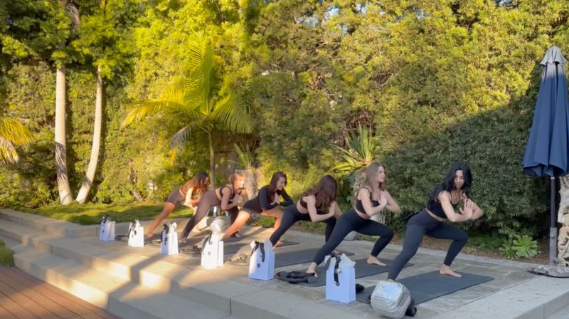 Sunlit outdoor yoga class on a garden patio — a group in black activewear doing lunge poses on mats by palm trees, white gift bags lined along the steps.