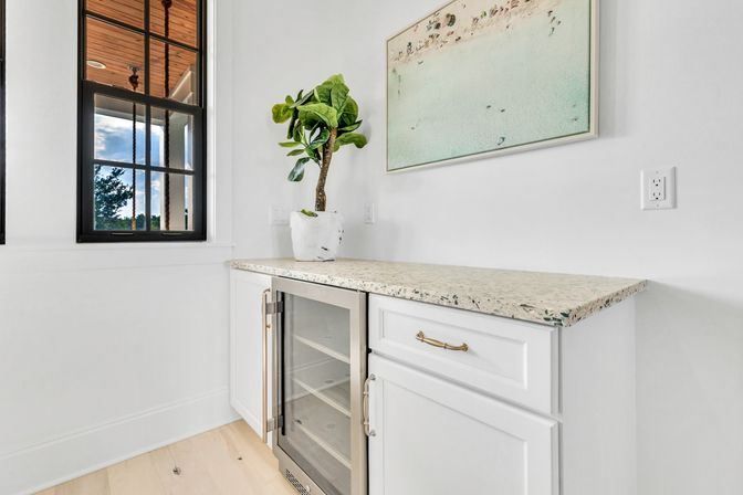 Bright modern kitchenette with white cabinetry, terrazzo countertop, built-in glass-front wine fridge, potted fiddle leaf fig, coastal wall art, black-framed window and light hardwood floor.