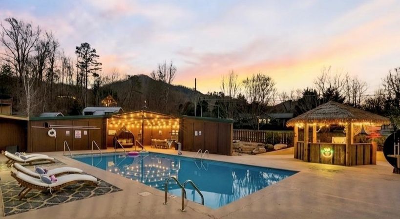 Outdoor mountain resort pool at dusk with string-lit wooden cabana and thatched tiki bar, lounge chairs on the deck and a pastel sunset reflecting in the water.