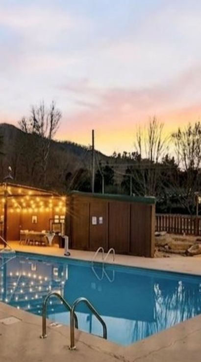 Outdoor mountain resort pool at dusk with string-lit wooden cabana and thatched tiki bar, lounge chairs on the deck and a pastel sunset reflecting in the water.