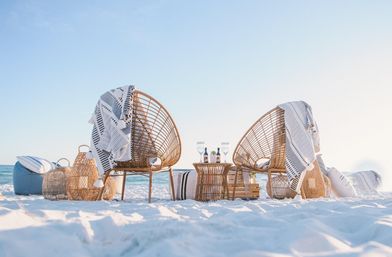 Cozy coastal setup: two wicker lounge chairs draped with striped throws on a white-sand beach facing the turquoise ocean, rattan table with wine glasses and woven baskets.