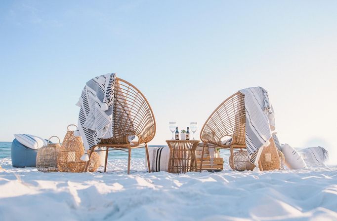 Cozy coastal setup: two wicker lounge chairs draped with striped throws on a white-sand beach facing the turquoise ocean, rattan table with wine glasses and woven baskets.