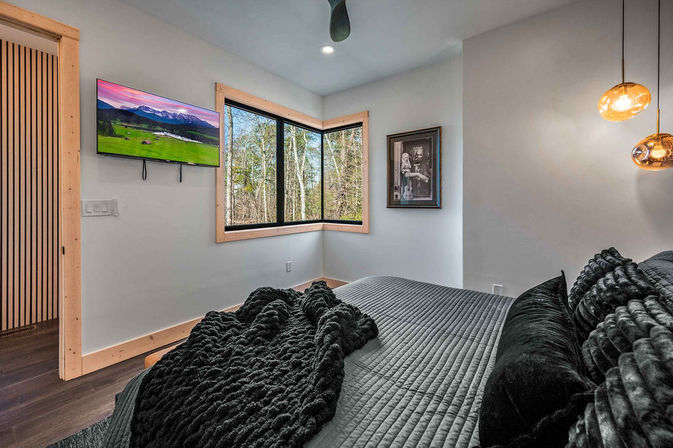 Cozy modern forest-view bedroom with gray quilted bed, plush black throw and velvet pillows, corner windows overlooking trees, wall-mounted TV, wood trim and amber pendant lights.