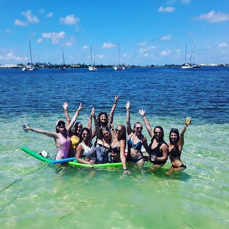 Group of friends in bikinis cheering in shallow turquoise-green coastal water with pool noodles and drinks, sailboats on the deep blue bay and a sunny blue sky on the horizon