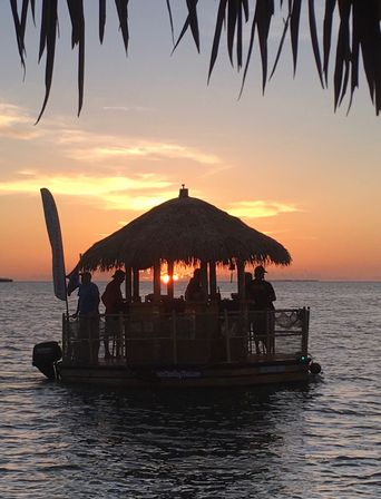 Silhouetted floating tiki bar raft framed by palm fronds, people enjoying a colorful tropical ocean sunset.