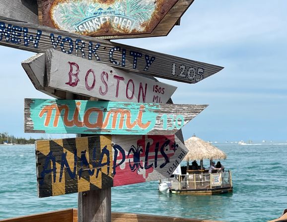 Colorful wooden directional signpost pointing to New York City, Boston, Miami and Annapolis, set against turquoise ocean waters and a thatched-roof tiki boat near a sunny pier