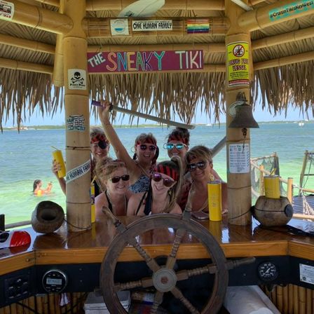 Group of friends in pirate bandanas and sunglasses posing behind a wooden ship wheel at an overwater tiki bar with a thatched roof, holding yellow cups and a prop sword, turquoise ocean and swimmers beyond.