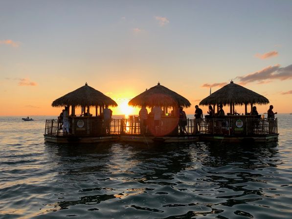 Silhouetted people on a floating tiki bar with three thatched huts at sunset, golden sky and sun reflecting on rippling ocean waters.