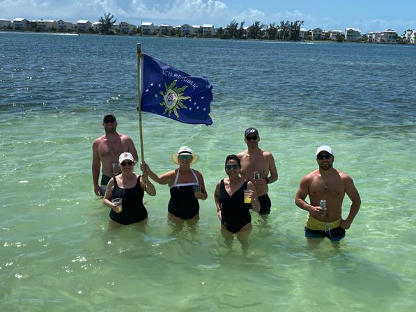 Six adults in swimsuits standing waist‑deep in clear turquoise coastal water, holding drinks and a blue flag with beachfront houses on the distant shore — sunny beach vacation vibe.
