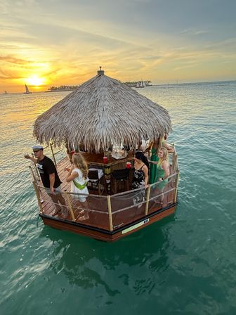 Floating tiki-bar raft with a thatched roof and people enjoying drinks on turquoise water at golden sunset, sailboats and a distant shoreline on the horizon.