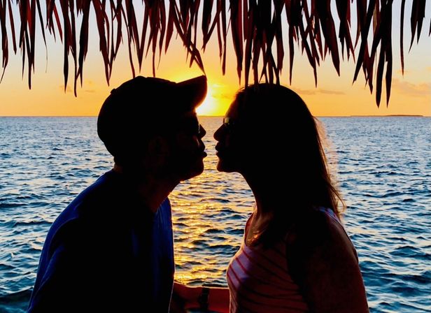 Silhouetted couple about to kiss beneath a thatched palm roof at a tropical ocean sunset, golden light reflecting on rippling water.
