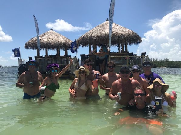 Group of people wading in clear turquoise shallow water at a sunny tropical sandbar, holding colorful drinks and posing in front of thatched tiki huts and a wooden dock under a blue sky