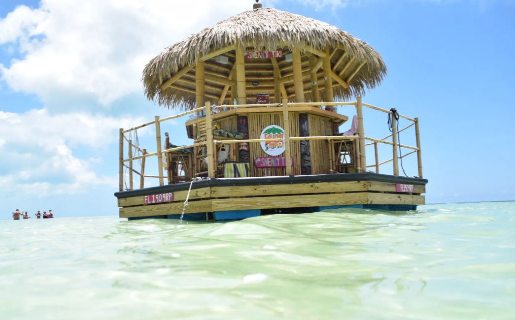 Floating thatched-roof tiki bar on a wooden raft in shallow turquoise ocean under a sunny blue sky, with people wading nearby — tropical vacation vibe.