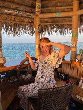 Person playfully shielding their eyes while steering a wooden ship wheel inside a thatched tiki hut overlooking calm ocean at sunset.