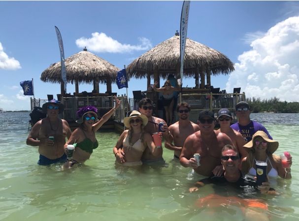 Group of friends enjoying drinks in shallow turquoise water in front of tiki huts on a sunny tropical sandbar, smiling and posing for a vacation photo.