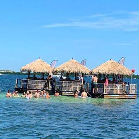 Floating tiki bar with three thatched-roof huts and people swimming and socializing in turquoise coastal waters under a bright blue sky
